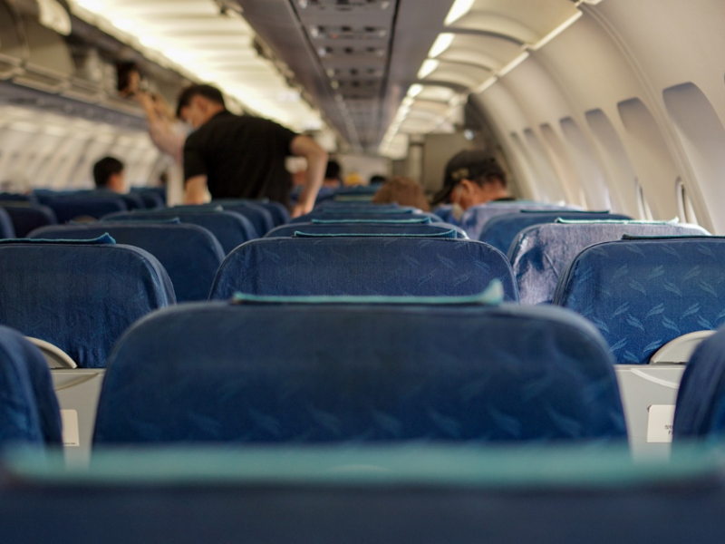 Eine Flugzeugkabine vom Sitz aus fotografiert. Einige Passagiere stehen von ihren Sitzen auf und holen ihr Gepäck aus der Gepäckablage des Flugzeugs.