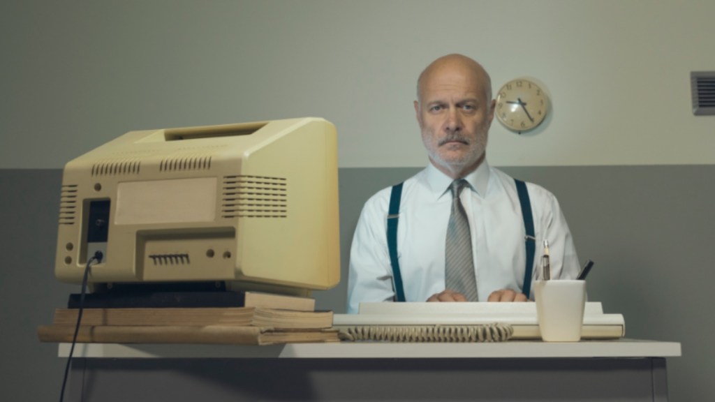 Älterer Geschäftsmann, der mit einem veralteten Computer an einem kleinen Schreibtisch arbeitet.